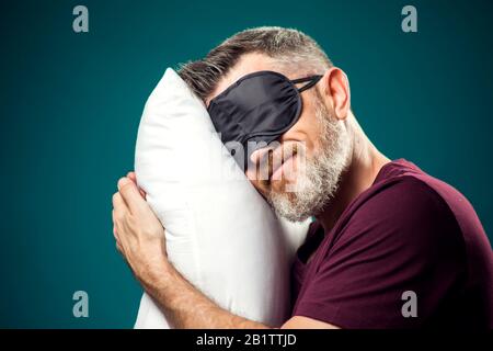 Mann in rotem T-Shirt und Schlafmaske auf dem Kopf, der weißes Kissen hält. Lifestyle- und Bettzeitkonzept Stockfoto