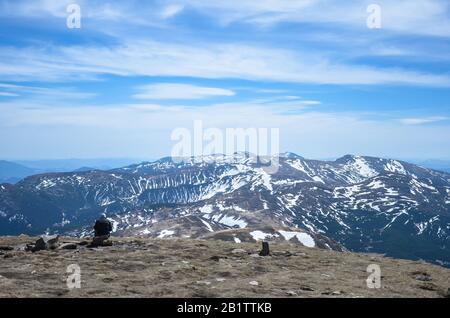 Man sitzt allein und betrachtet die schönen schneereichen Berggipfel am sonnigen Tag, Karpatengebirge, Ukraine. Man Traveler sitzt auf Felsen Stockfoto