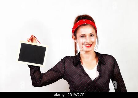 Junge kaukasische fröhliche Rotkopffrau mit roten Lippen und Bandana, die schwarze leere Tafel hält und lächelt Stockfoto