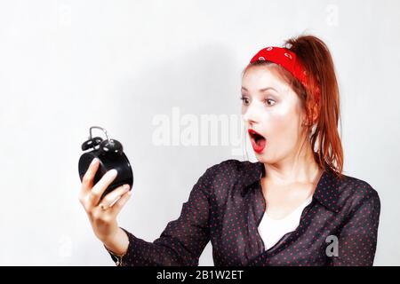 Junge lächelnde kaukasische Ansteckerin mit roten Lippen und Bandana, die peinlich aussieht und Angst vor schwarzen Metall-Vintage-Uhren hat, die in der Hand halten Stockfoto