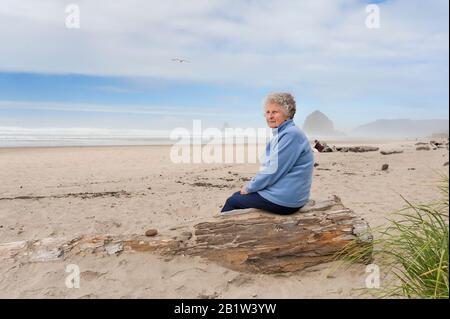 Cannon Beach, Oregon, USA - 28. September 2010: Eine ältere Frau sitzt auf einem Stück Treibholz am Cannon Beach an der Oregon Coast. Stockfoto