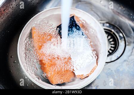 Wasser gießt Waschmittel in der Küche Edelstahlwaschbecken mit Kunststoffschale, die zwei Stücke frischen bio-organischen Lachses enthält Stockfoto