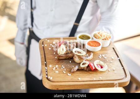 Der Kellner hält einen Teller Lamb Zungenkebab mit Eingelegtem Kohl und zwei Soßen. Serviert auf einem Holzbrett. Grillrestaurant, eine Serie Stockfoto