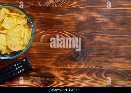 Kartoffelchips in einer Glasschale neben der TV-Fernbedienung auf einem gefeuerten Holzhintergrund mit Kopierraum. Draufsicht. Nahaufnahme. Stockfoto