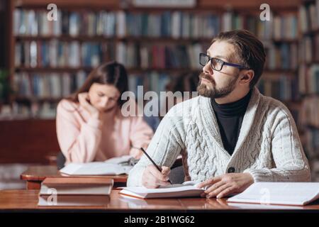 Nachdenklicher bärtiger Mann im Polokragen und Strickpullover am Tisch sitzen und Informationen in Büchern suchen. Reifer Mann, der Zeit in der öffentlichen waage verbringt Stockfoto