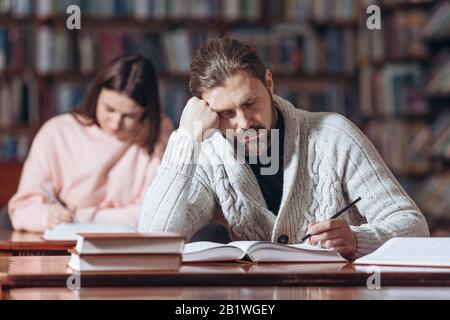 Müde bärtige Männer in schwarzem Polokragen und Strickpullover schlafen ein, während sie an der öffentlichen Universität nach notwendigen Informationen suchen. Reifer Leser schläfrft ein Stockfoto
