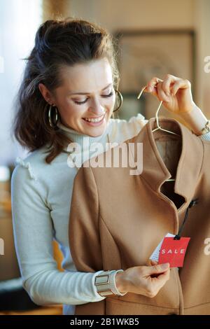 Lächelnde elegante Frau in weißem Pullover und Rock im modernen Modegeschäft untersucht den Verkaufspreis auf beigefarbenem Mantel. Stockfoto