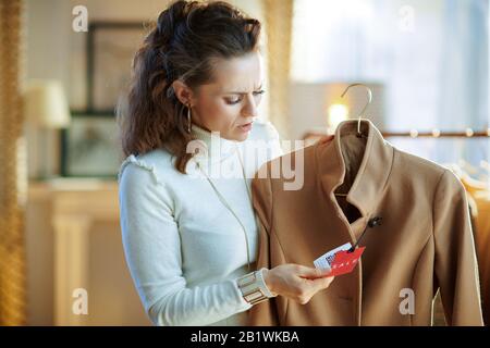 Besorgt elegante Frau in weißem Pullover und Rock im modernen Mode-Showroom untersucht den Verkaufspreis auf beigefarbenem Mantel. Stockfoto
