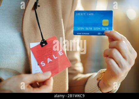 Nahaufnahme der Frau im weißen Pullover mit beigefarbenem Mantel mit rotem Kaufpreisschild und blauer Kreditkarte in der modernen Modeboutique. Stockfoto