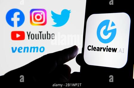 Das Logo des Unternehmens Clearview AI auf dem Smartphone hält sich mit unscharfen sozialen Medien in den Händen, in denen die Daten erfasst wurden. Konzept für die Gesichtserkennungssoftware. Stockfoto