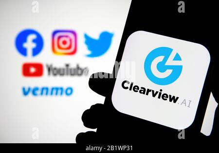 Das Logo des Unternehmens Clearview AI auf dem Smartphone hält sich mit unscharfen sozialen Medien in den Händen, in denen die Daten erfasst wurden. Konzept für die Gesichtserkennungssoftware. Stockfoto