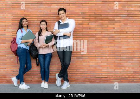 Drei asiatische lächelnde Studenten, die Buch und Laptop auf Backstein-Hintergrund im Campus halten. Gruppe glücklicher Schüler der Teenager im Freien. Stockfoto