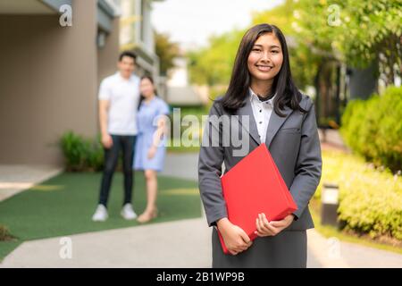 Asiatische selbstbewusste Frau Immobilienmaklerin oder Immobilienmaklerin im Anzug, die rote Feile hält und mit jungen Paar-Hausverkäufern hinter sich vor dem Haus lächelt. Portrai Stockfoto