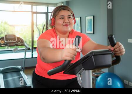 Übergewichtige asiatische Frau mit Ohrhörertraining auf dem Heimtrainer in der modernen Sporthalle, glücklich und lächelnd beim Workout. FAT Woman kümmert sich um Gesundheit und Stockfoto