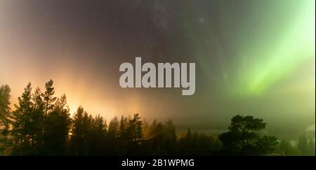 Panoramablick auf die leuchtend grüne Aurora, die über die schwedische Nebelwaldlandschaft scheint, Lichtstrahlen von einem Dorf und Nordlicht-Farbhimmel in der Luft Stockfoto