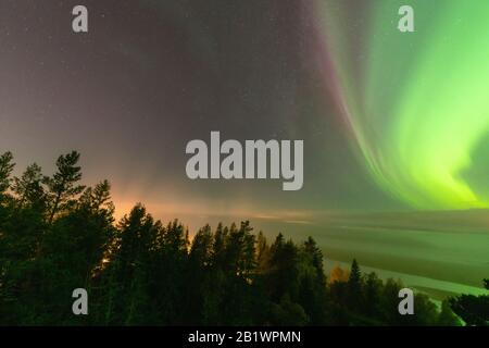 Blick auf die leuchtend grüne Aurora, die über die schwedische Nebelwaldlandschaft in den Bergen glänzt, Lichtstrahlen aus einem Dorf und Nordlichter färben Himmel in di Stockfoto