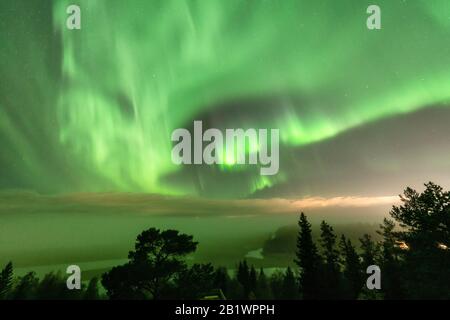 Blick auf die leuchtend grüne Aurora, die über die schwedische Nebelwaldlandschaft in den Bergen glänzt, Lichtstrahlen aus einem Dorf und Nordlichter färben Himmel in di Stockfoto