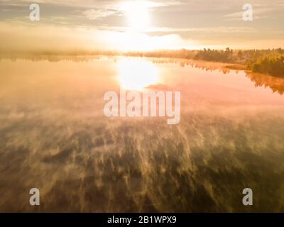 Schöne Luftaufnahme über Sommer See mit Nebel über Wasseroberfläche und Sonnenaufgang über Waldlandschaft und Dorf, still Luft. Rot orange Sonne Stockfoto