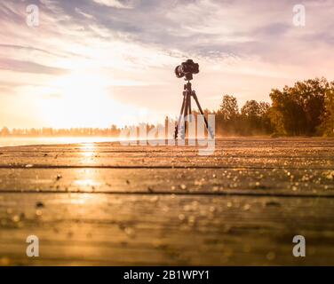 Stativfotokamera auf Holzbrückenfilmen foggy hellen Sonnenaufgang über See und Bäumen Stockfoto