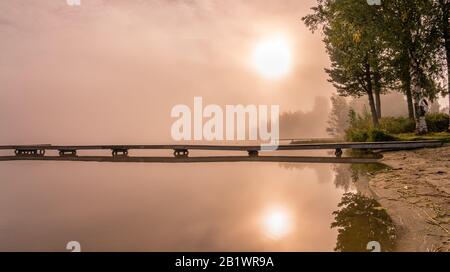 Wunderschöne goldene Sonne leuchtet durch Nebel am See während des frühen Sonnenaufgangs, lange Holzbrücke, Bäume, Himmel Reflexion in ruhigem Wasser, kopieren Raum Stockfoto