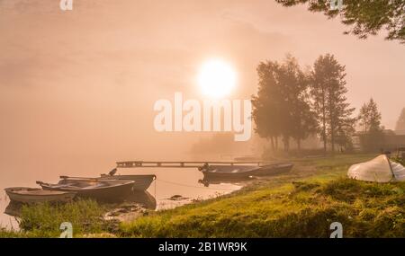 Wunderschöne goldene Sonne leuchtet durch Nebel am See während des frühen Sonnenaufgangs, Boote und lange Holzbrücke, Bäume, Himmel Reflexion in ruhigem Wasser, Copy Space Stockfoto