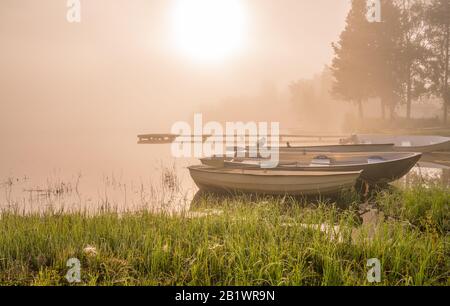 Schöne goldene Sonne leuchtet bei frühem Sonnenaufgang durch Nebel am See, Boote und lange Holzbrücke, Bäume, Himmelsreflexion in ruhigem Wasser Stockfoto