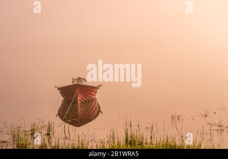 Ein kleines Motorboot, schöne goldene Sonne leuchtet bei frühem Sonnenaufgang durch dichten Nebel am See, ruhiges Wasser Stockfoto