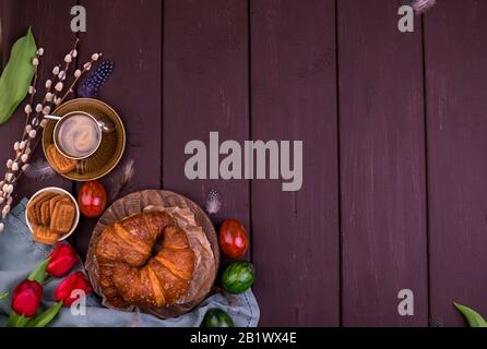 Osterfrühstück mit aromatischem frischem Espresso, Croissants, farbigen Eiern, roten Tulpen und Weide. Kaffee mit Gebäck, Blumen auf einem Holztisch. Ansicht von oben. Kopierbereich Stockfoto