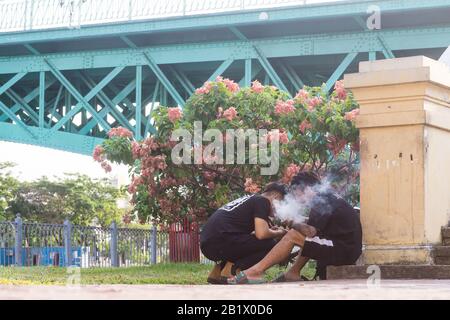 Saigon (Ho-Chi-Minh-Stadt)-Straßenszene - zwei vietnamesische Männer in ihren Zwanzigern rauchen und spielen auf ihren Telefonen unterhalb der Mong-Brücke in Saigon. Stockfoto