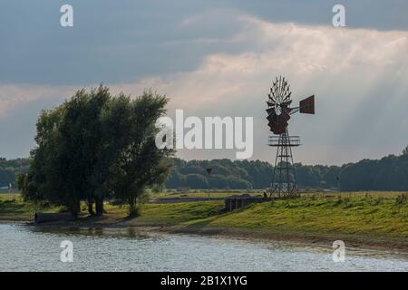 Bäume und Bauernwindmühle am Ufer einer Auenwiese neben einem Flussbett mit Jakobs Leiter-Sonnenstrahlen im Hintergrund Stockfoto