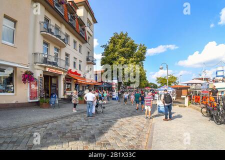 Touristen genießen die lebhaften bunten Geschäfte und Cafés an der Hauptpromenade des Alter Strom Kanals entlang der Ostsee in Warnemunde Deutschland. Stockfoto