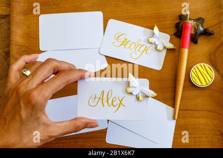 Hochzeitskalligraphie - weibliche Hand mit handschriftlichen Platzkarten mit Namen und Schmetterlingen aus Papier. Goldene Schrift auf Whitepaper. Stockfoto