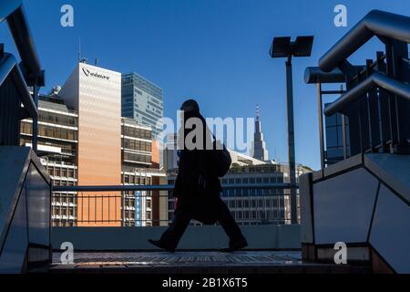 Ein japanischer Salaryman oder Büroangestellter geht auf einer Fußgängerbrücke in der Nähe des Shinjuku-Bahnhofs mit dem Docomo-Turm dahinter. Shinjuku, Tokio, Japan Stockfoto