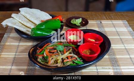 Tongericht mit Rinderfajitas mit Pfeffer- und Zwiebelstreifen, begleitet von einem Teller mit Maistortillas und würziger Soße und Guacamol auf einem Holztisch Stockfoto