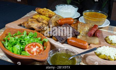 Großes Holztablett mit gegrilltem Fleisch und Würstchen, begleitet von Soßen, gebratenem Maniok, einem Teller mit Reis und Tomaten und Salat auf einem schwarzen Tisch Stockfoto