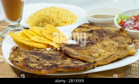 Weiße Platte mit Schweinekopf, begleitet von gebratener Banane, Salat und gelbem Reis mit einer Glaskasse mit Limonade auf weißem Hintergrund Stockfoto