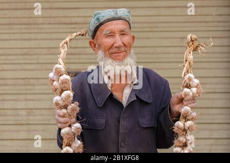 Älterer uyghur-mann mit grauem Bart, der Knoblauchschnüre an einem Basar in Kaschgar hält. Der Mann trägt einen traditionellen Doppelhut. Stockfoto