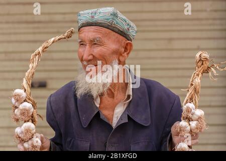 Lächelnder, älterer uyghur-mann mit einem Doppelkopf, der zwei Knoblauchstränge auf einem Markt in Kaschgar hält. Stockfoto