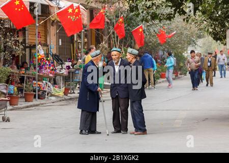 Drei ältere Männer der Minderheit der Uyghur haben ein Gespräch auf einer Straße in der Kaschgar-Altstadt. Chinesische Flaggen sind hinten auf dem Haus angebracht. Stockfoto