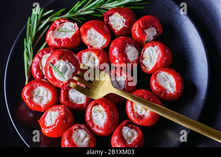 Vegetarische Appetithappen rote Kirschpfeffer gefüllt mit Frischkäse auf schwarzem Teller mit Rosmarinsprig auf dunklem Betongrund, Nahaufnahme Stockfoto