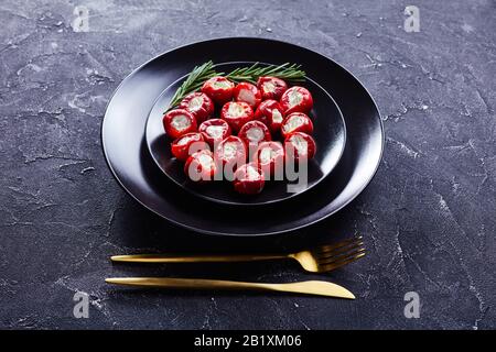 Pikante Kirschpfeffer mit weichem Ricotta-Käse auf schwarzem Teller mit Rosmarinsprigen mit goldenem Besteck auf dunklem Betongrund, Stockfoto