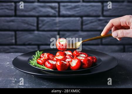 Frau hält goldene Gabel mit Kirschpfeffer, gefüllt mit weichem Ricotta-Käse, serviert auf einem schwarzen Teller mit Rosmarinsprig auf einem dunklen Betonbac Stockfoto