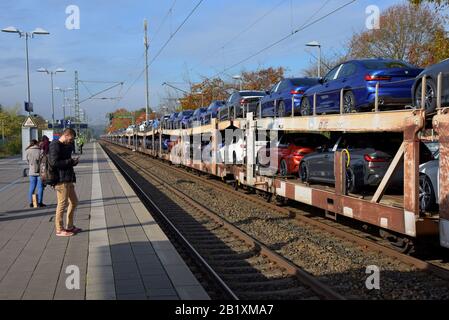 Passagiere, die auf einen Zug der Deutschen Bahn im Bahnhof Buxtehude warten, während ein Güterzug, der neue BMW-Wagen transportiert, durchfährt. Oktober 2019 Stockfoto