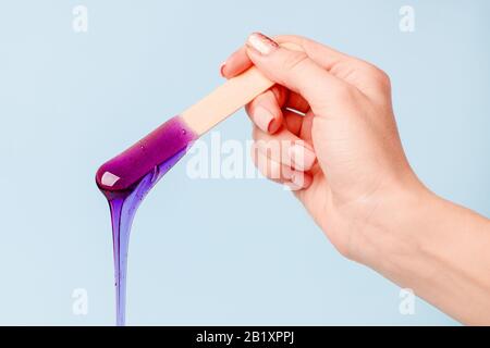 Hand hält violettes Wachs oder Zuckerpaste auf Holzspachtel, die auf blauem Grund in den Behälter hinabfließen. Konzept der Werbeschönheitsbranche, l. Stockfoto