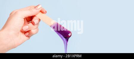 Hand hält violettes Wachs oder Zuckerpaste auf Holzspachtel, die auf blauem Grund in den Behälter hinabfließen. Konzept der Werbeschönheitsbranche, l. Stockfoto