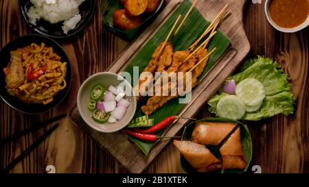 Blick auf thailändische traditionelle Straßennahrung mit Schweinesatay mit Erdnusssauce, thailändischen Würstchen, Khao Soi und gebratenen Frühlingsrollen auf rustikalem Tisch Stockfoto