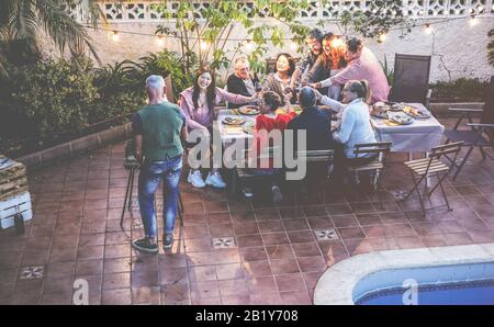 Fröhliche Familie, die beim Barbecue-Party-Dinner mit Rotwein isst und jubelt - Verschiedene Altersgruppen, die Spaß am grillmenü haben, sitzen im Hinterhof der Villa - S Stockfoto