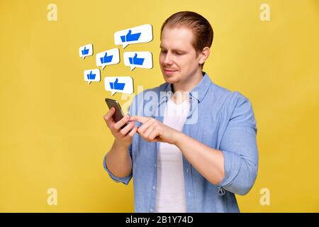 Ein Mann in einem Hemd auf gelbem Hintergrund sieht wie in einem Smartphone aus. Abstrakte Symbole mit dem Daumen nach oben in der Luft. Nahaufnahme. Stockfoto