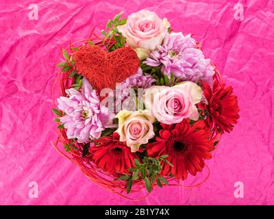 Roter und rosafarbener Blumenstrauß mit Rosen, gerberblüten, Dahlien und herzförmigem Schmuck auf rosa Hintergrund Stockfoto