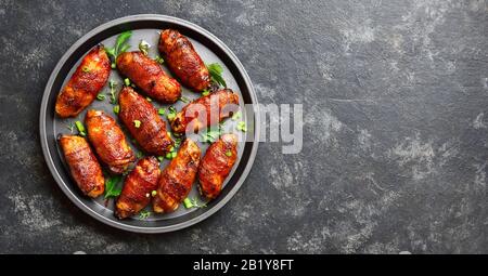 Speck umwickelt gegrillte Chicken Wings auf die Platte über der schwarzen Stein Hintergrund mit freier Platz für Text. Leckeren snack von Hühnerfleisch, Speck in Süß, sauer Stockfoto